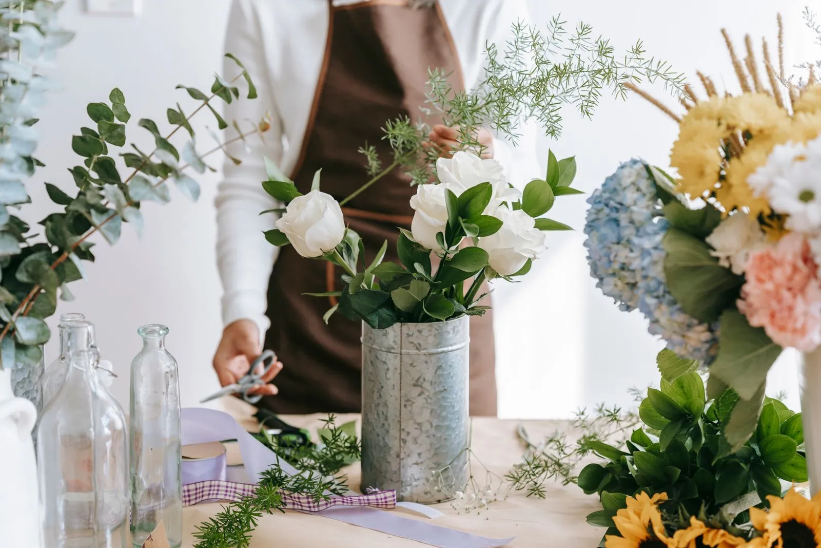 Crop unrecognizable person with Asparagus plant sprig against blooming roses and Ruscus in vase on table in shop